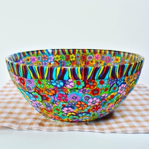 May include: A colourful decorative bowl with a floral design. The bowl is covered in small, brightly coloured flowers in shades of pink, blue, yellow, and orange. The rim has a striped pattern. The bowl sits on a checkered cloth.