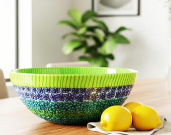 May include: A ceramic bowl with a vibrant floral design. The bowl features a green rim with vertical stripes, a band of blue and purple flowers, and a green floral pattern. Three yellow lemons rest on a white cloth on a wooden table.