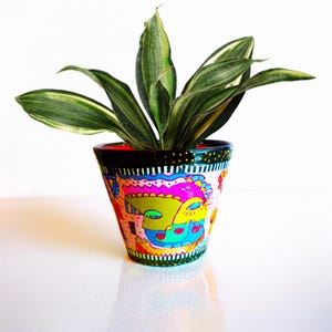 Pode incluir: Vaso de flores de cerâmica pintado à mão com um design abstrato colorido. O vaso apresenta uma borda preta com pontos dourados, um design de rosto rosa e azul e uma planta listrada verde e branca. Contém uma planta verde.