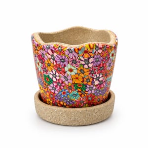May include: A flower pot with a wavy rim and a matching saucer, both featuring a speckled beige texture. The exterior of the pot is covered in a dense, colorful pattern of small, multi-colored flowers in shades of pink, orange, yellow, and blue.