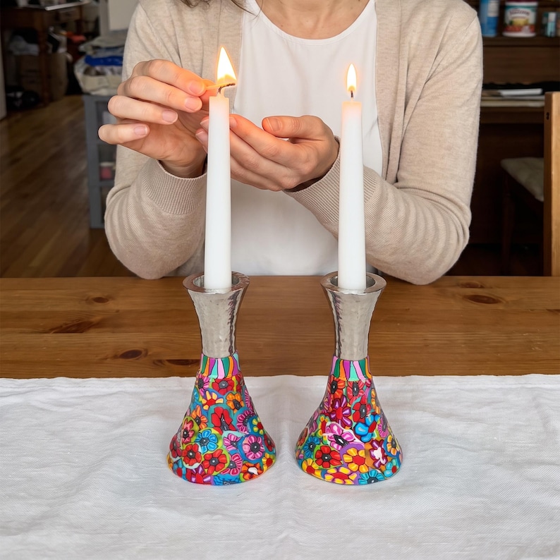 May include: Two tall white taper candles standing in matching metal candle holders. The base of each holder features a vibrant, multi-colored floral pattern with red, pink, yellow, and blue petals. The upper section of the holders is silver-toned.