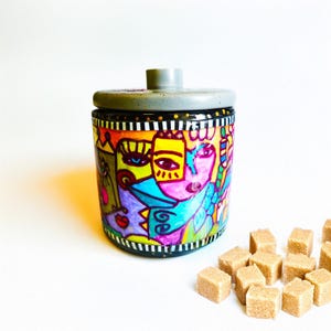 May include: A colorful ceramic jar with a gray lid, featuring a hand-painted design of abstract faces in vibrant hues. The jar is surrounded by brown sugar cubes, suggesting a kitchen or dining theme.