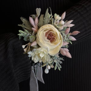 May include: A white and pink flower boutonniere with greenery and white berries. The boutonniere is tied with a gray ribbon.
