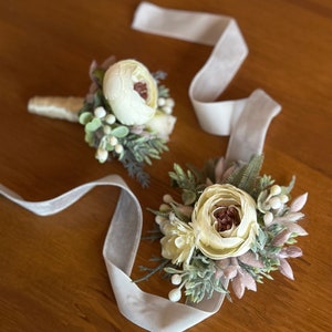 May include: Two white flower boutonnieres with greenery and a white satin ribbon. The boutonnieres are perfect for a wedding or special occasion.