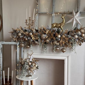 May include: A festive mantelpiece decorated with gold and silver ornaments, including a golden reindeer figurine and a white star. The garland features gold leaves, silver berries, and hanging ornaments. A small table holds a floral arrangement with a reindeer figurine.