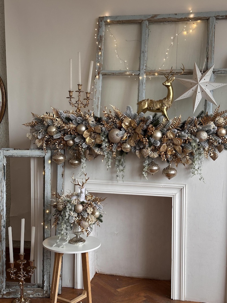 May include: A festive mantelpiece decorated with gold and silver ornaments, including a golden reindeer figurine and a white star. The garland features gold leaves, silver berries, and hanging ornaments. A small table holds a floral arrangement with a reindeer figurine.