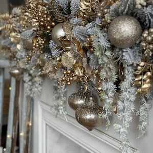 May include: A festive Christmas garland with gold and silver ornaments and frosted greenery. The garland features gold-painted leaves, berries, and several glittery ball ornaments. The arrangement is set against a white background, creating a luxurious holiday display.