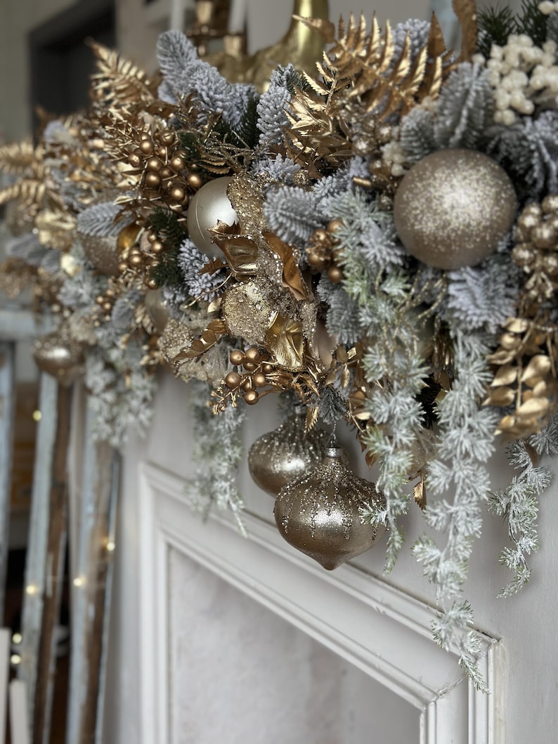 May include: A festive Christmas garland with gold and silver ornaments and frosted greenery. The garland features gold-painted leaves, berries, and several glittery ball ornaments. The arrangement is set against a white background, creating a luxurious holiday display.