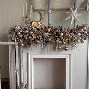 May include: A festive Christmas mantelpiece decorated with gold and silver ornaments, faux greenery, and a golden reindeer figurine. The mantel also features a candelabra with white candles and a string of fairy lights, creating a warm, inviting holiday display.