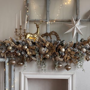 May include: A festive Christmas mantel arrangement featuring a garland of gold and silver foliage, gold ornaments, and a golden reindeer figurine. The display includes white paper star decorations and a candelabra.