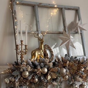 May include: A festive Christmas mantel display featuring a gold reindeer figurine, a gold candelabra with white candles, and gold ornaments. The display includes a garland of gold and silver foliage, and string lights. Two white paper star decorations are also visible.