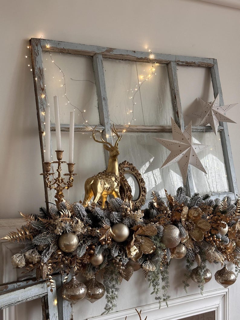 May include: A festive Christmas mantel display featuring a gold reindeer figurine, a gold candelabra with white candles, and gold ornaments. The display includes a garland of gold and silver foliage, and string lights. Two white paper star decorations are also visible.