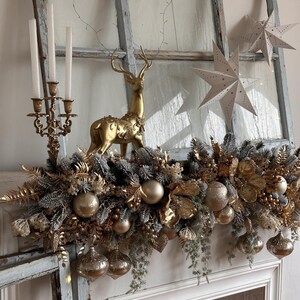May include: A festive Christmas mantel arrangement featuring a gold reindeer figurine, a gold candelabra with white candles, and hanging ornaments. The arrangement includes gold and silver decorative elements, including star-shaped decorations and a garland.