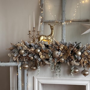 May include: A festive Christmas mantelpiece decoration featuring a gold reindeer figurine, gold ornaments, and faux greenery. The arrangement includes gold-colored baubles, leaves, and berries, creating a luxurious holiday display. The mantel is decorated with string lights.