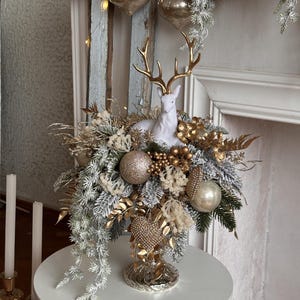May include: A festive centerpiece featuring a white ceramic deer with gold antlers, surrounded by gold and silver ornaments, faux greenery, and decorative elements. The arrangement is displayed on a white table, creating a holiday theme.