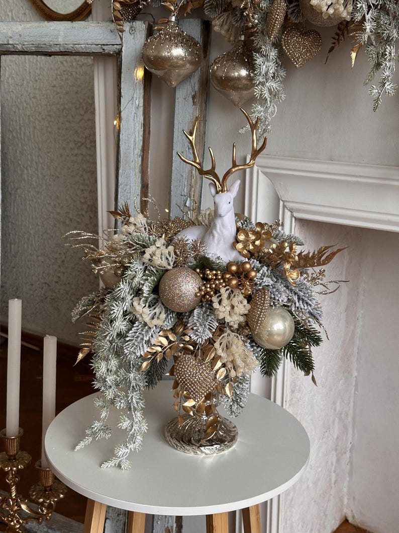 May include: A festive centerpiece featuring a white ceramic deer with gold antlers, surrounded by gold and silver ornaments, faux greenery, and decorative elements. The arrangement is displayed on a white table, creating a holiday theme.
