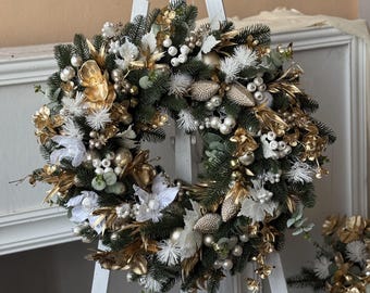 May include: A festive Christmas wreath with a circular design. The wreath features green pine needles, white flowers, gold leaves, and silver and gold ornaments. It is displayed on a white easel.