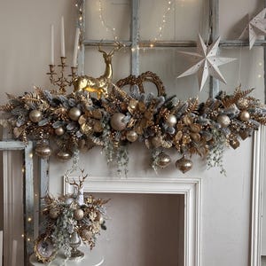 May include: A festive Christmas mantelpiece arrangement featuring gold and silver ornaments, faux foliage, and a golden reindeer figurine. The display includes hanging ornaments, decorative stars, and a candelabra.