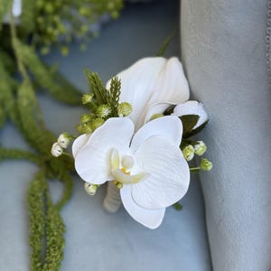 May include: A boutonniere featuring a white orchid blossom as the focal point, accented with small green sprigs, tiny white buds, and dark green leaves. The floral arrangement is secured with a wrapped stem base, creating a compact design.