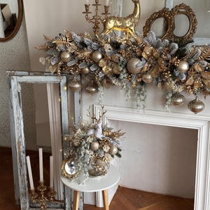 May include: A festive Christmas mantel arrangement featuring gold and silver ornaments, faux greenery, and a golden reindeer figurine. The display includes a garland, decorative spheres, and a small table with a floral arrangement. A vintage mirror and candelabras add to the holiday decor.