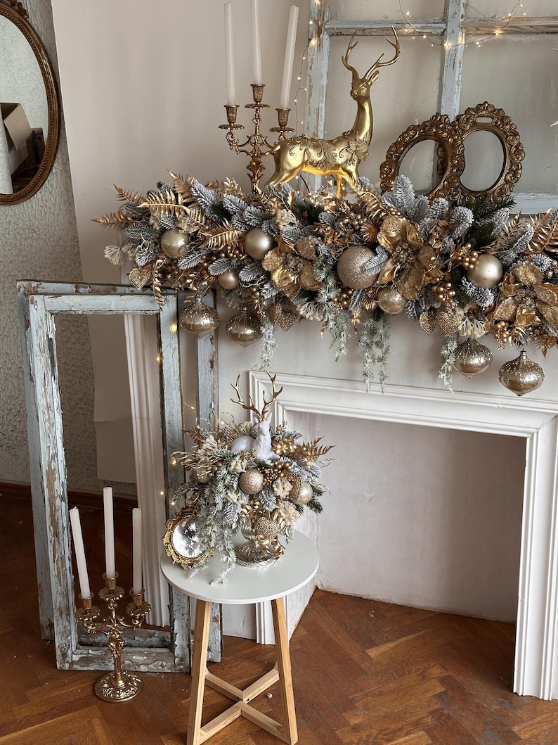 May include: A festive Christmas mantel arrangement featuring gold and silver ornaments, faux greenery, and a golden reindeer figurine. The display includes a garland, decorative spheres, and a small table with a floral arrangement. A vintage mirror and candelabras add to the holiday decor.