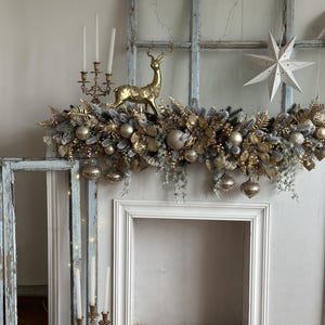 May include: A festive Christmas mantelpiece decorated with a gold and silver garland, ornaments, and a golden reindeer figurine. A white star-shaped decoration hangs to the right. Antique candle holders and a distressed window frame add to the holiday decor.