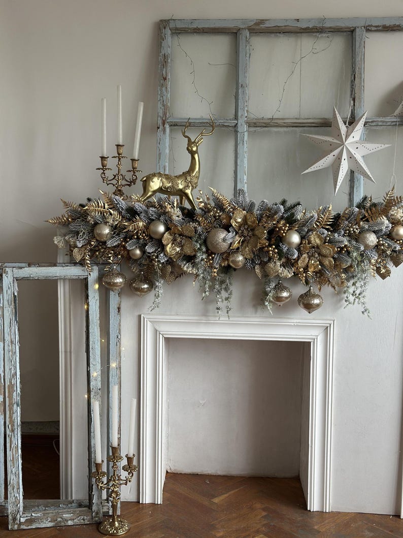 May include: A festive Christmas mantelpiece decorated with a gold and silver garland, ornaments, and a golden reindeer figurine. A white star-shaped decoration hangs to the right. Antique candle holders and a distressed window frame add to the holiday decor.
