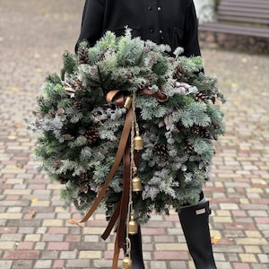 Op de afbeelding: Een grote, ronde kerstkrans gemaakt van groenblijvende takken, dennenappels en bevroren accenten. Bruine linten en kleine gouden belletjes hangen aan de krans. De krans wordt vastgehouden door een persoon die een zwarte jas en zwarte laarzen draagt.