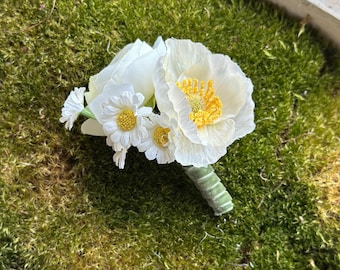 Puede incluir: Un prendido floral blanco con una gran flor tipo amapola con centro amarillo, rodeada de flores más pequeñas tipo margarita. Los tallos están envueltos en tela verde. El arreglo descansa sobre un lecho de musgo verde.