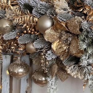 May include: A festive Christmas wreath featuring gold and silver ornaments, faux frosted pine needles, and gold floral accents. The wreath includes several gold-colored baubles and decorative elements, creating a luxurious holiday display.