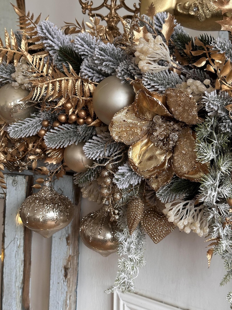 May include: A festive Christmas wreath featuring gold and silver ornaments, faux frosted pine needles, and gold floral accents. The wreath includes several gold-colored baubles and decorative elements, creating a luxurious holiday display.