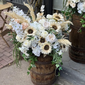 Può includere: Una grande composizione floreale con girasoli, rose, ortensie e piume di erba della pampa color crema e bianco. I fiori sono esposti in una rustica botte di legno marrone scuro con fasce metalliche, arricchita da edera verde e fogliame pendente.