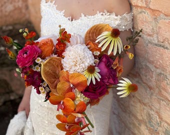 Puede incluir: Un ramo de flores en cascada con orquídeas de color naranja vibrante, ranúnculos magenta intenso, dalias blancas y equináceas de color amarillo pálido. El arreglo incluye varias flores en tonos otoñales y cintas largas de color naranja y rosa.