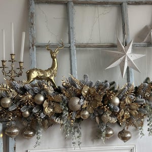 May include: A festive Christmas garland with gold and silver ornaments, faux greenery, and a golden reindeer figurine. The garland is adorned with gold baubles and hanging ornaments. Two white paper star decorations are in the background. A gold candelabra with three candles is also visible.