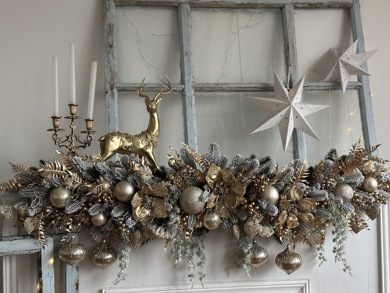 May include: A festive Christmas garland with gold and silver ornaments, faux greenery, and a golden reindeer figurine. The garland is adorned with gold baubles and hanging ornaments. Two white paper star decorations are in the background. A gold candelabra with three candles is also visible.