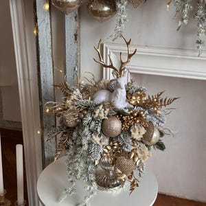 May include: A festive Christmas centerpiece featuring a white ceramic deer figurine with gold antlers, surrounded by gold and silver ornaments, faux greenery, and decorative accents. The arrangement is displayed on a white table, creating a holiday atmosphere.
