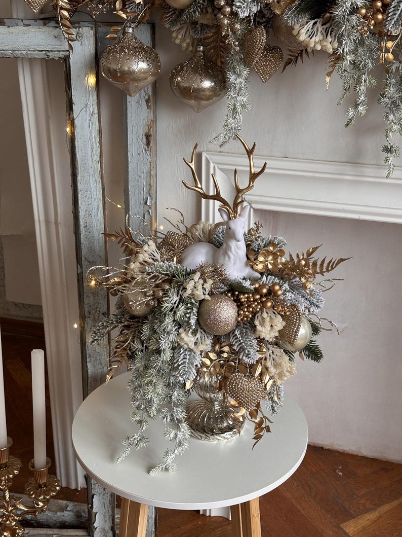 May include: A festive Christmas centerpiece featuring a white ceramic deer figurine with gold antlers, surrounded by gold and silver ornaments, faux greenery, and decorative accents. The arrangement is displayed on a white table, creating a holiday atmosphere.