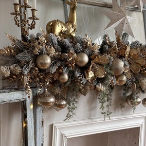 May include: A festive Christmas mantel decoration featuring a gold reindeer figurine, gold ornaments, and a garland of gold and silver foliage. The arrangement includes a gold candelabra with white candles and star-shaped paper ornaments.