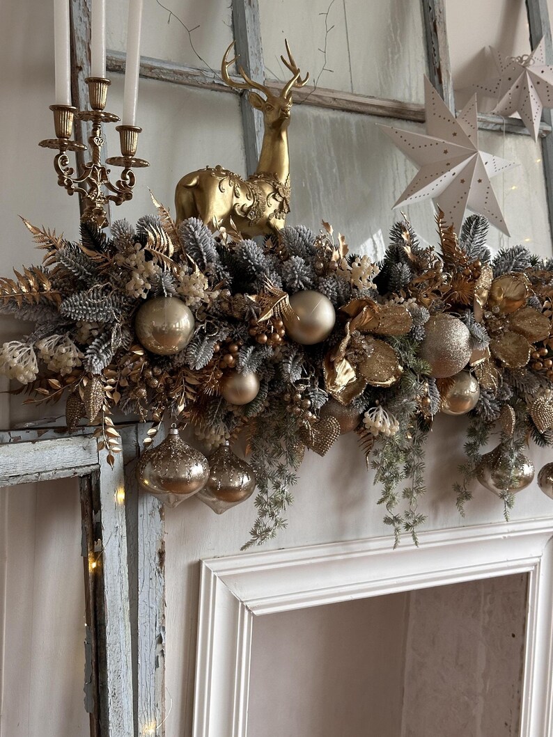 May include: A festive Christmas mantel decoration featuring a gold reindeer figurine, gold ornaments, and a garland of gold and silver foliage. The arrangement includes a gold candelabra with white candles and star-shaped paper ornaments.