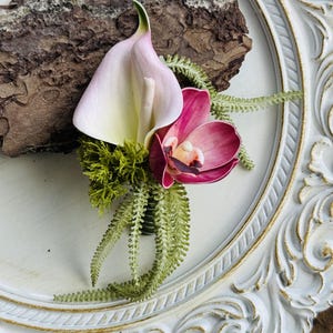 Puede incluir: Un prendedor floral con un lirio de cala blanco y rosa pálido, una orquídea magenta intenso, musgo verde y delicadas hojas de helecho. El arreglo está colocado sobre un trozo de corteza de árbol marrón texturizada y un marco blanco adornado.