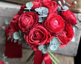 May include: A floral bouquet featuring deep red roses and ranunculus flowers interspersed with pale green eucalyptus leaves. The stems are gathered together and tied with a long, smooth red satin ribbon that hangs down from the base of the flowers.