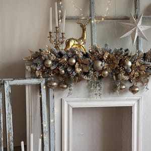 May include: A festive Christmas mantelpiece decorated with gold and silver ornaments, faux greenery, and a golden reindeer figurine. The mantel is framed by a white fireplace and an antique window with string lights. A white star and candles complete the holiday display.