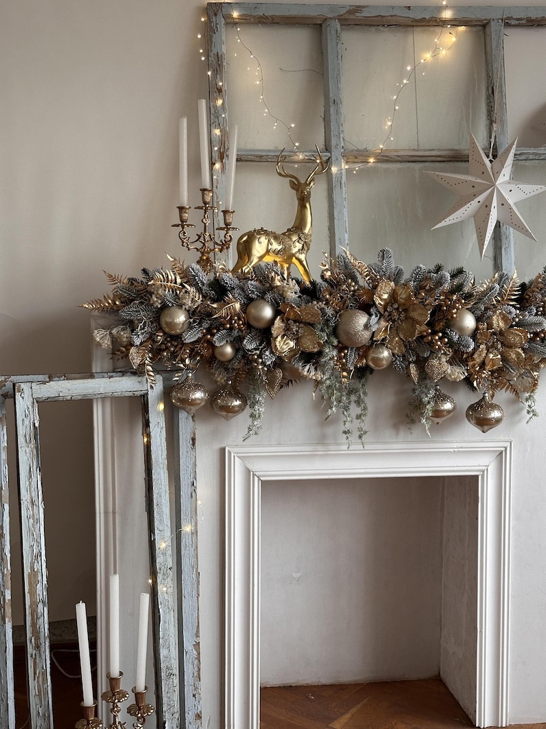 May include: A festive Christmas mantelpiece decorated with gold and silver ornaments, faux greenery, and a golden reindeer figurine. The mantel is framed by a white fireplace and an antique window with string lights. A white star and candles complete the holiday display.