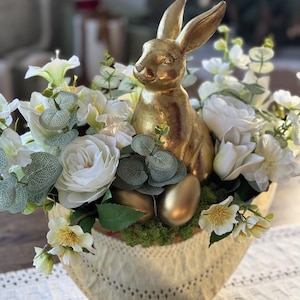 Könnte beinhalten: Eine goldfarbene Hasenfigur sitzt inmitten weißer Rosen, Grünzeug und goldener Eier in einem dekorativen Korb. Der Korb hat eine cremefarbene, strukturierte Außenseite. Dieses Arrangement zum Thema Ostern eignet sich für die Inneneinrichtung.