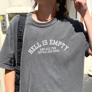 May include: A charcoal gray, short-sleeved t-shirt with the text "HELL IS EMPTY AND ALL THE DEVILS ARE HERE" in white. The shirt has a slightly faded appearance and a crew neck. The word "pepper" is in the bottom right corner.