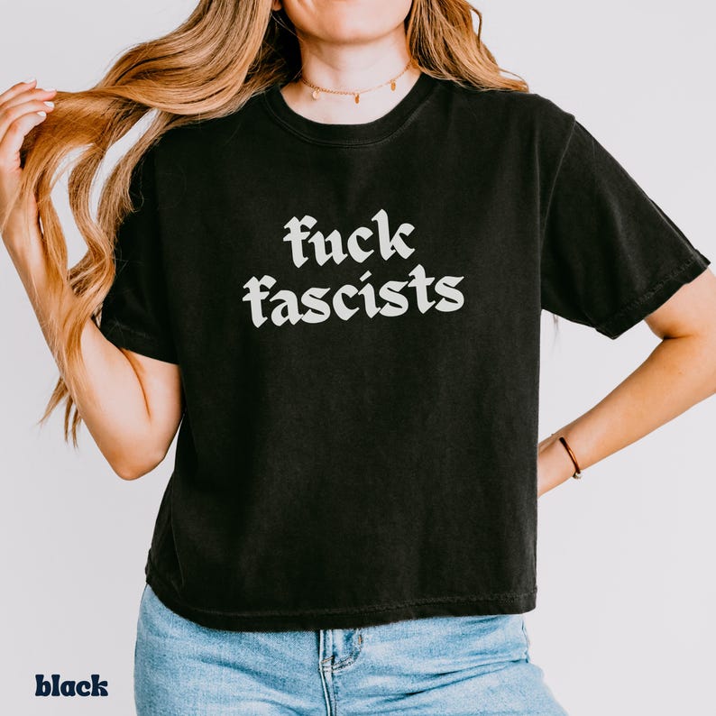 May include: Black t-shirt with the text "fuck fascists" in a white gothic font. The shirt is a crew neck style and appears to be made of a soft material. The word "black" is printed at the bottom left of the image.