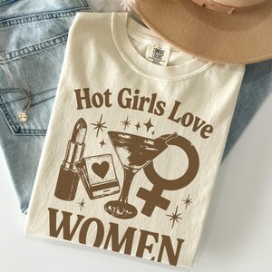 May include: Cream-colored t-shirt with the text "Hot Girls Love WOMEN" in brown. The graphic includes a lipstick, a matchbook with a heart, a martini glass, and a female symbol. The shirt is folded on top of blue jeans.