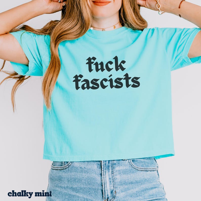 May include: A light blue t-shirt with the words "fuck fascists" in black, gothic-style font. The shirt is a chalky mint color and is paired with blue jeans. The words "chalky mint" are at the bottom left of the image.