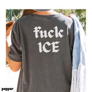 May include: Dark gray t-shirt with the words "fuck ICE" in white gothic font. The shirt is a short-sleeved style. A denim jacket with fringe is draped over the shoulder.