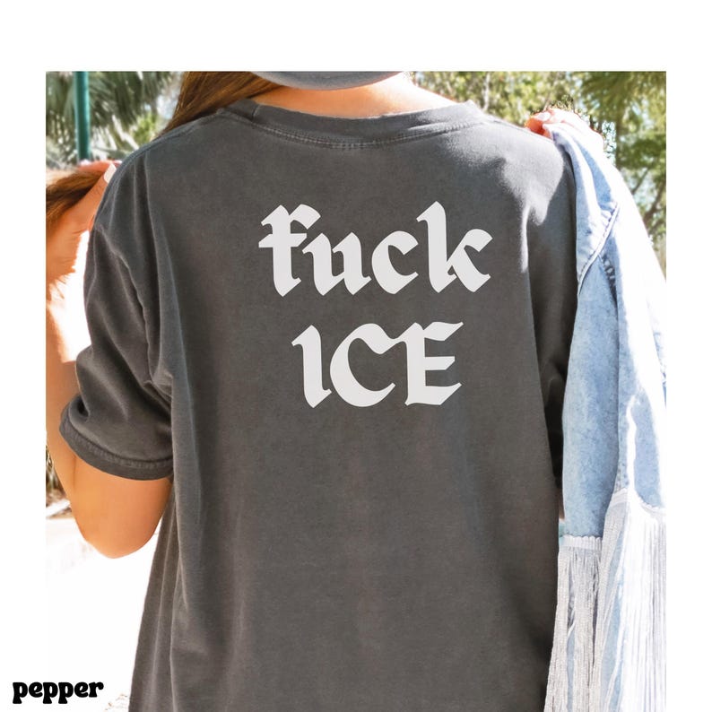 May include: Dark gray t-shirt with the words "fuck ICE" in white gothic font. The shirt is a short-sleeved style. A denim jacket with fringe is draped over the shoulder.
